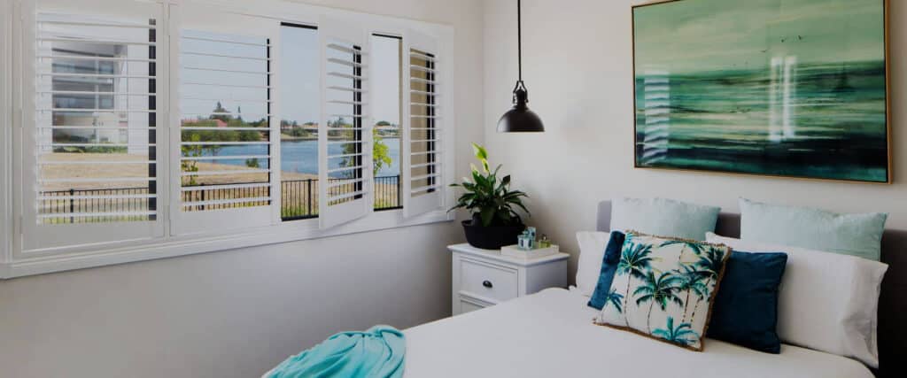 White indoor shutters in bedroom showing bed with white bedspread and tropical themed pillows and picture above