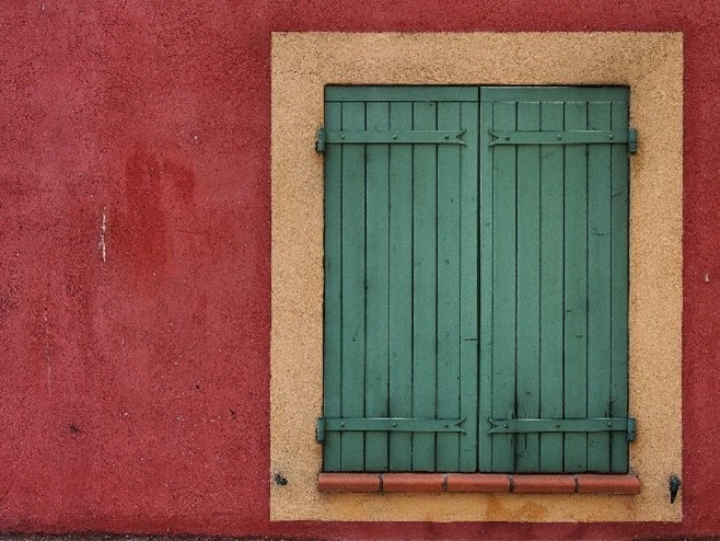 closed green wooden shutters covering a yellow window opening on a red building wall